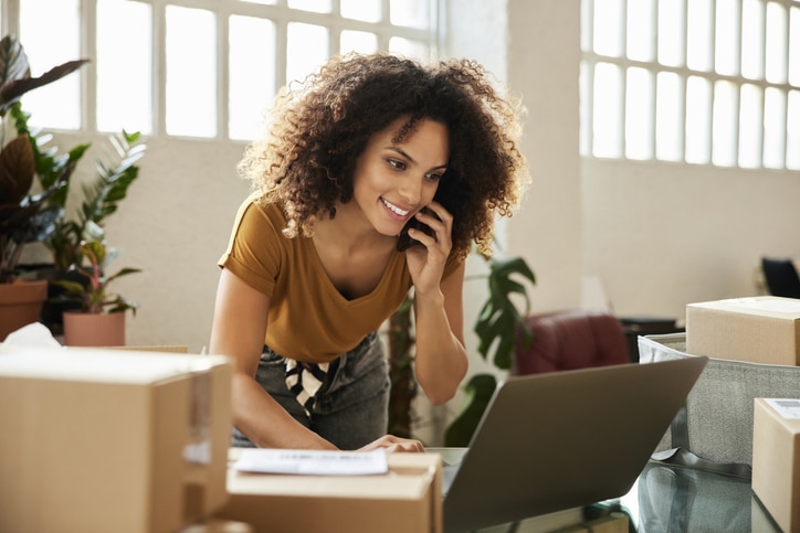 Young female talking on smart phone while using laptop. Confident businesswoman is working at home office. She has curly hair.